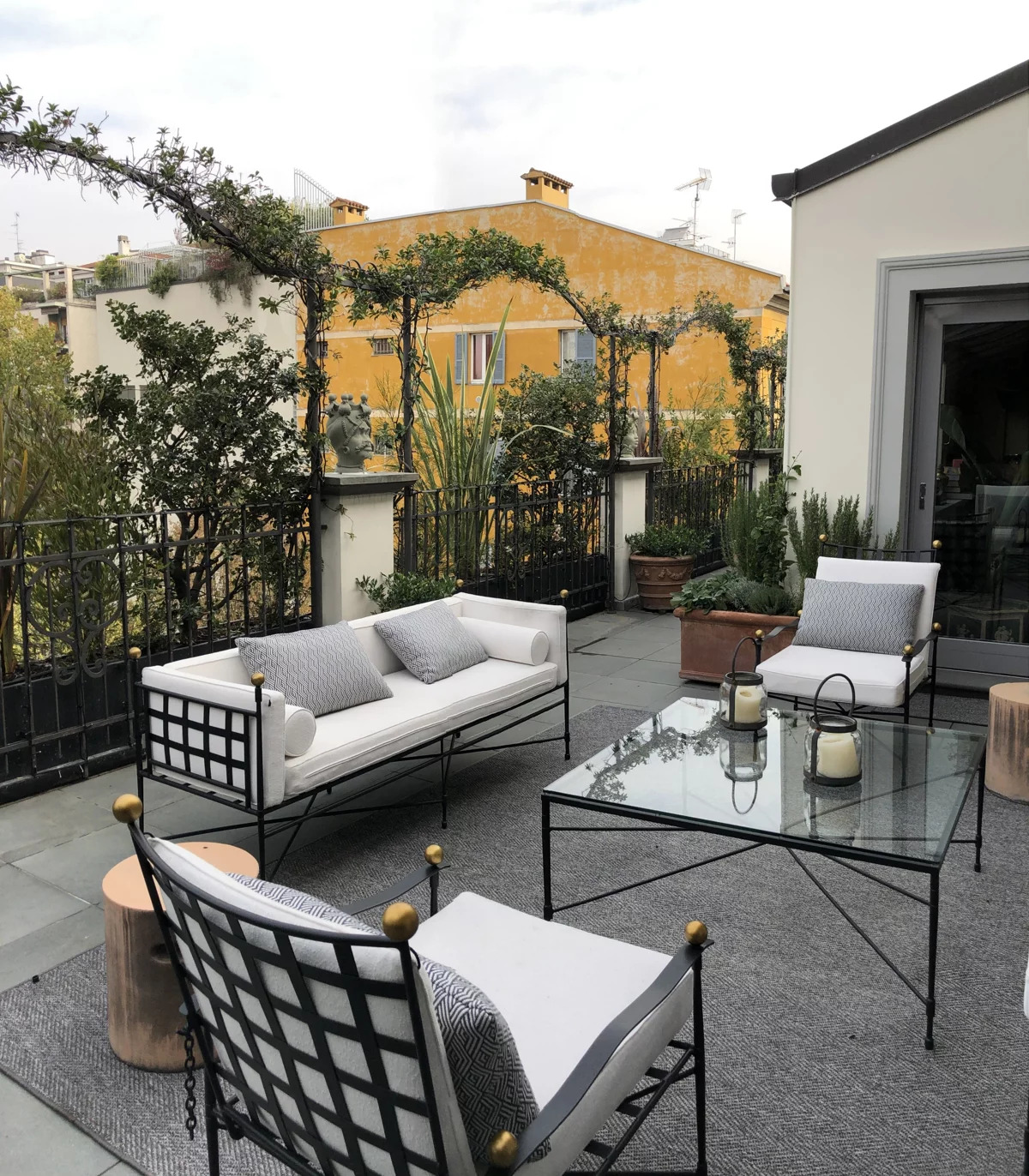 Terrazzo con divano e poltrone in metallo, tavolino in vetro e pergolato di rampicanti davanti a una facciata gialla